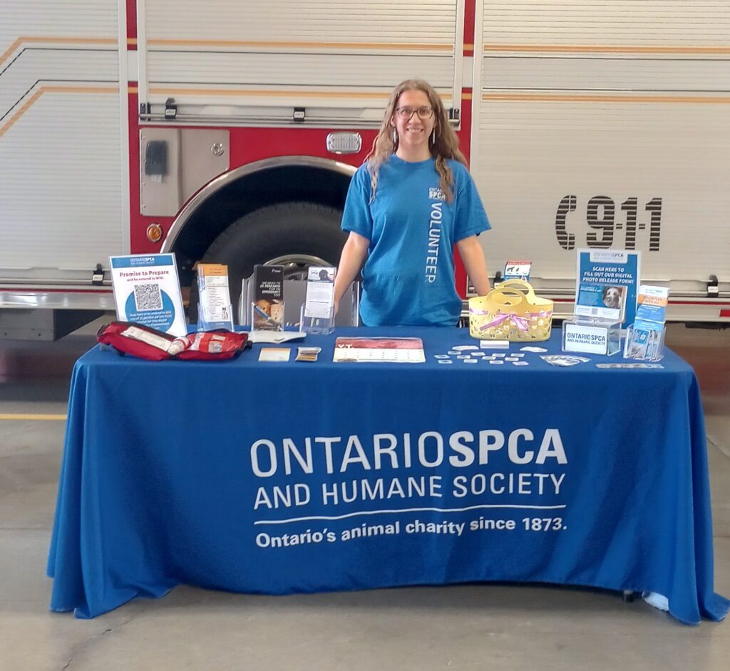 Ontario SPCA and Humane Society volunteer stands behind an informational display table covered in brochures and promotional items, set up in front of a fire truck at a community event.
