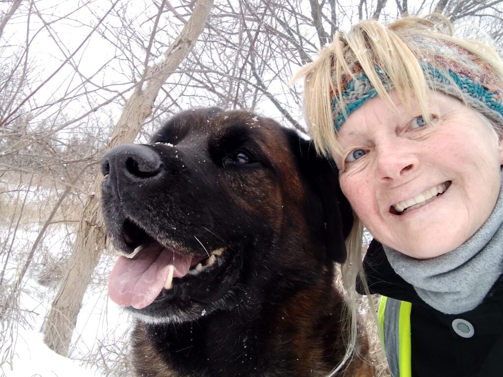 Smiling woman in winter clothing takes a selfie with a large black and brown dog on a snowy trail surrounded by bare trees.