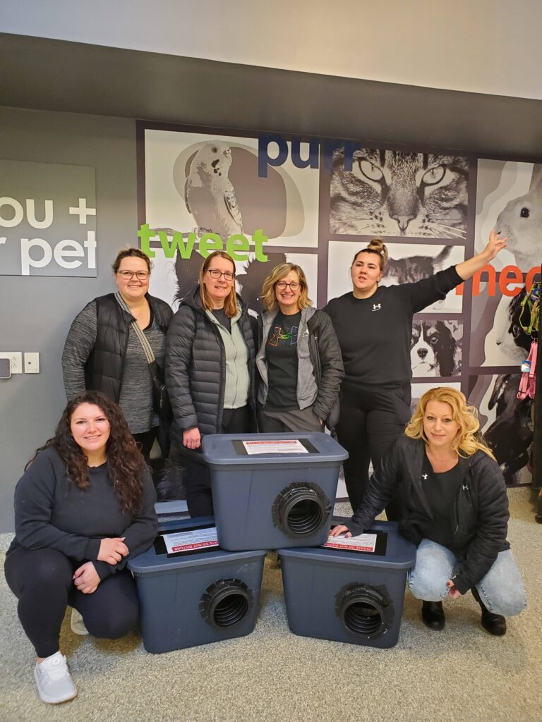 Group of six people posing with assembled outdoor cat shelters inside an animal centre
