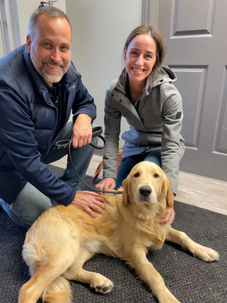 Smiling man and woman kneel beside a relaxed Golden Retriever lying on a black mat inside a room with grey walls and doors.