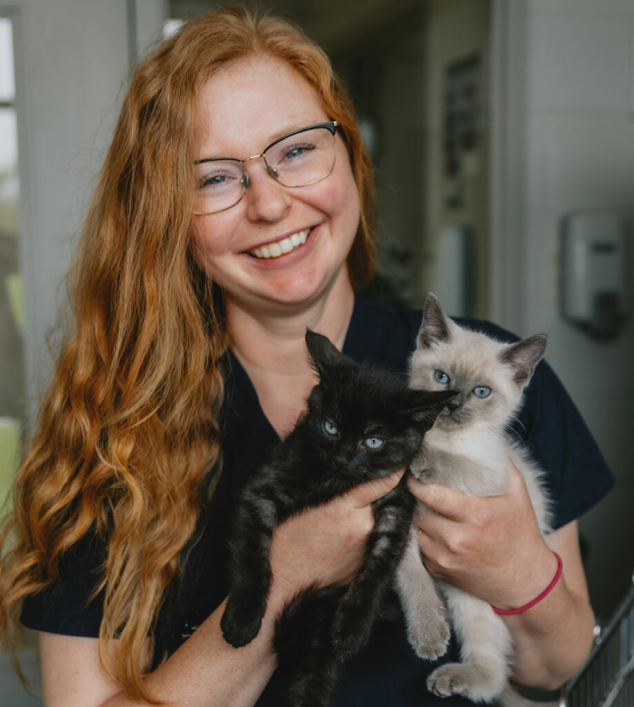 Smiling woman with long red hair and glasses holds two kittens—one black and one cream with grey markings—inside an animal care facility.