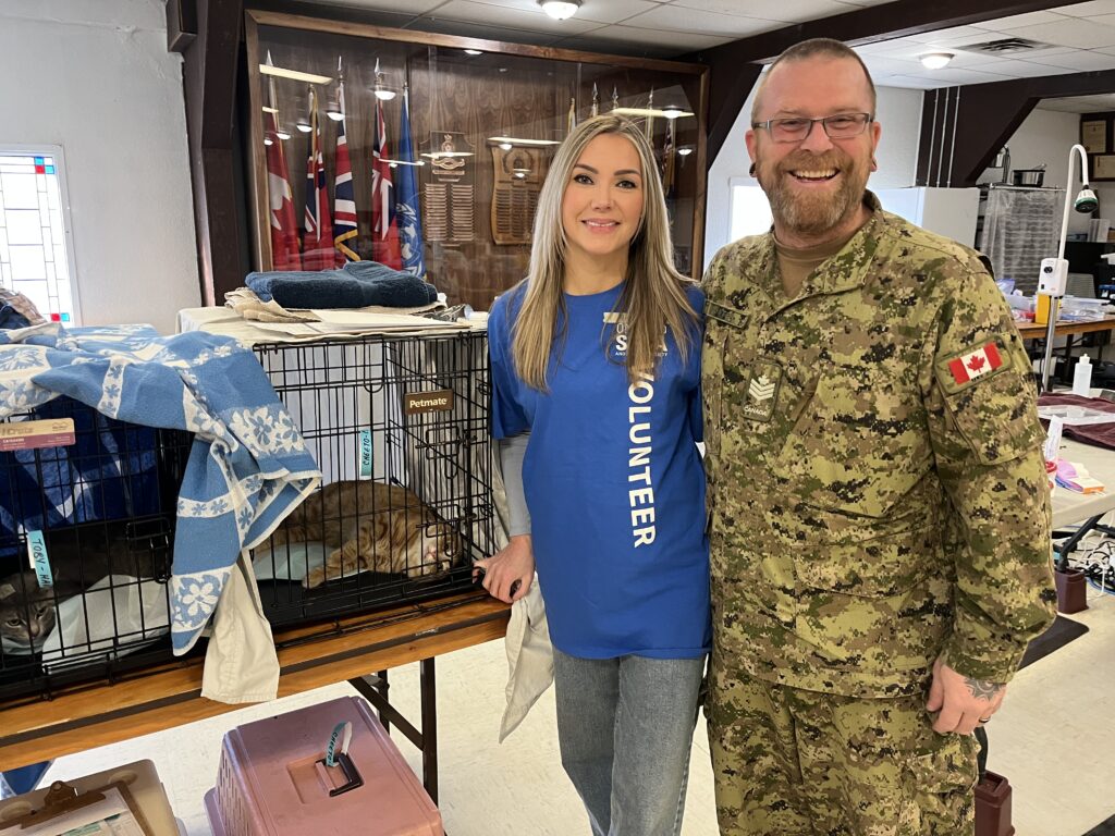 Smiling woman in a blue volunteer shirt stands beside a man in Canadian military uniform, next to a kennel covered with blankets inside a community hall.