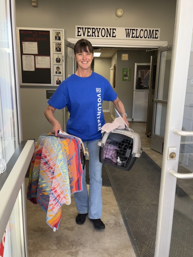 Smiling Ontario SPCA volunteer wearing a blue shirt carries two covered pet carriers while walking through the entrance of a community building with a sign that reads “Everyone Welcome.”