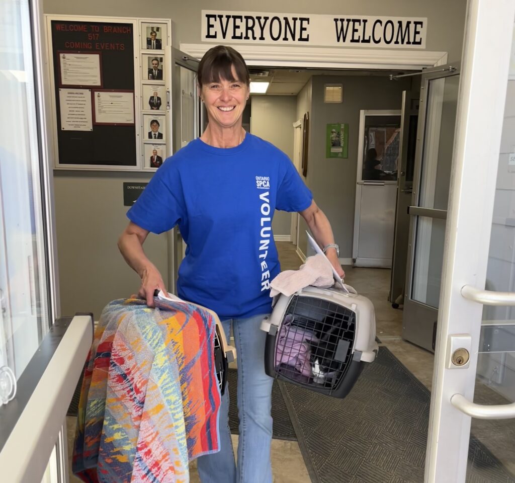 Smiling Ontario SPCA volunteer wearing a blue shirt carries two covered pet carriers while walking through the entrance of a community building with a sign that reads “Everyone Welcome.”