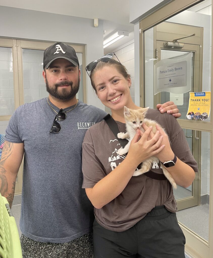 A smiling man and woman stand indoors, the woman holding a small orange and white kitten.