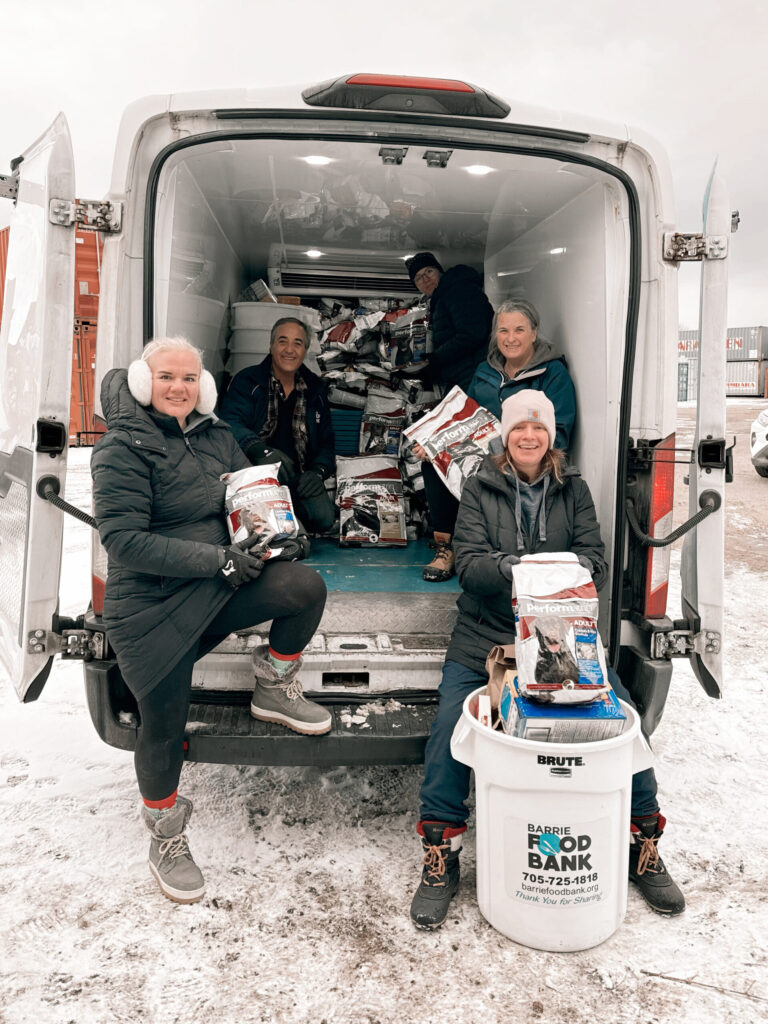 Group of five people dressed in winter clothing sit and stand at the back of a van filled with bags of pet food, with a Barrie Food Bank donation bin in front on a snowy day.