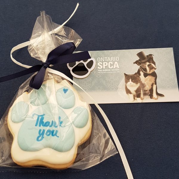 Paw-shaped cookie with blue icing and 'Thank you' text, wrapped in clear plastic with a navy ribbon, alongside an Ontario SPCA wedding-themed card featuring a dog in a top hat and a cat