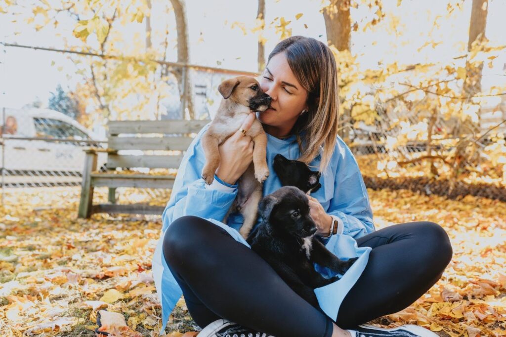 A woman sitting on the ground, smiling while holding three playful puppies in her arms.