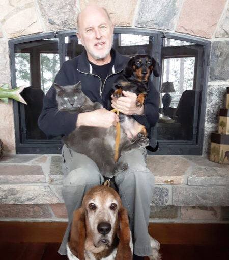 Man sitting in front of a stone fireplace holding a grey cat and a small black-and-tan Dachshund, with a Basset Hound sitting at his feet.