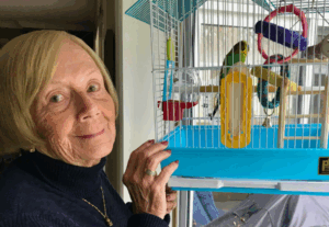 Judie standing beside a blue birdcage, smiling at her two green and yellow budgies inside.