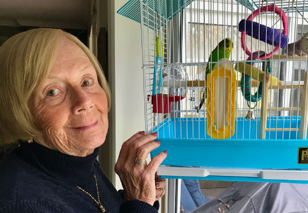 Judie standing beside a blue birdcage, smiling at her two green and yellow budgies inside.