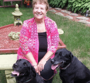 Linda sitting outdoors on a garden bench, smiling beside her two black Labrador Retrievers.