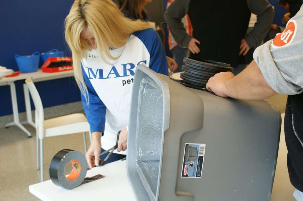 Person cuts duct tape beside a grey storage bin being made into an insulated feral cat shelter