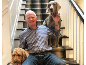 John sitting on a staircase smiling with two dogs — a golden doodle and a Weimaraner — beside him.