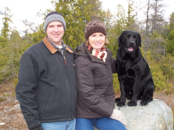 Couple sitting outdoors in winter clothing with their black Labrador retriever on a rock, surrounded by evergreen trees.