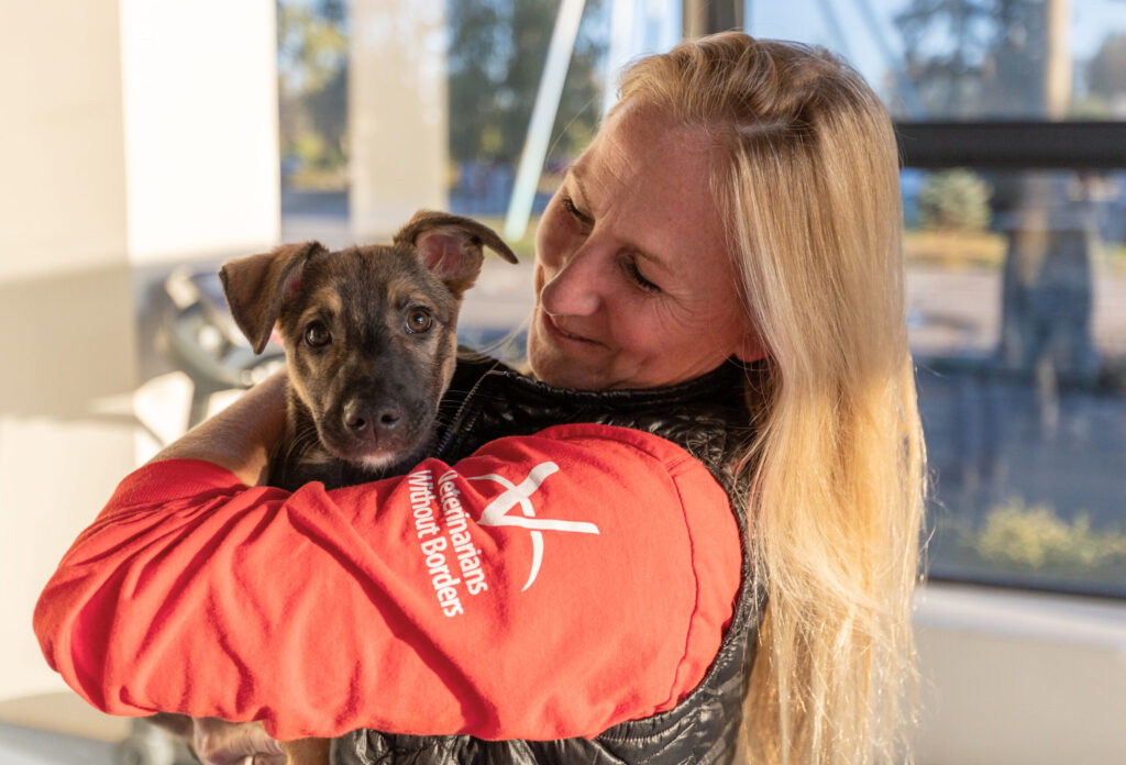 A woman in a red Veterinarians Without Borders shirt smiling and holding a brown puppy in her arms.