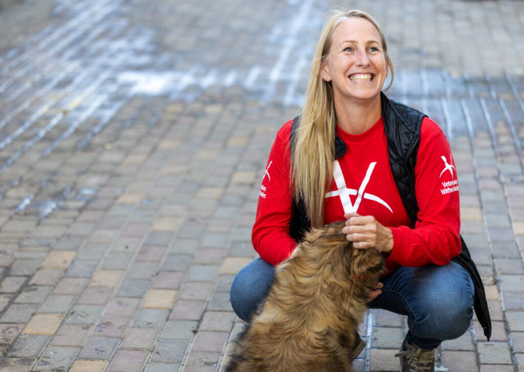 A smiling woman in a red Veterinarians Without Borders shirt kneeling and petting a brown dog outdoors on a brick walkway.
