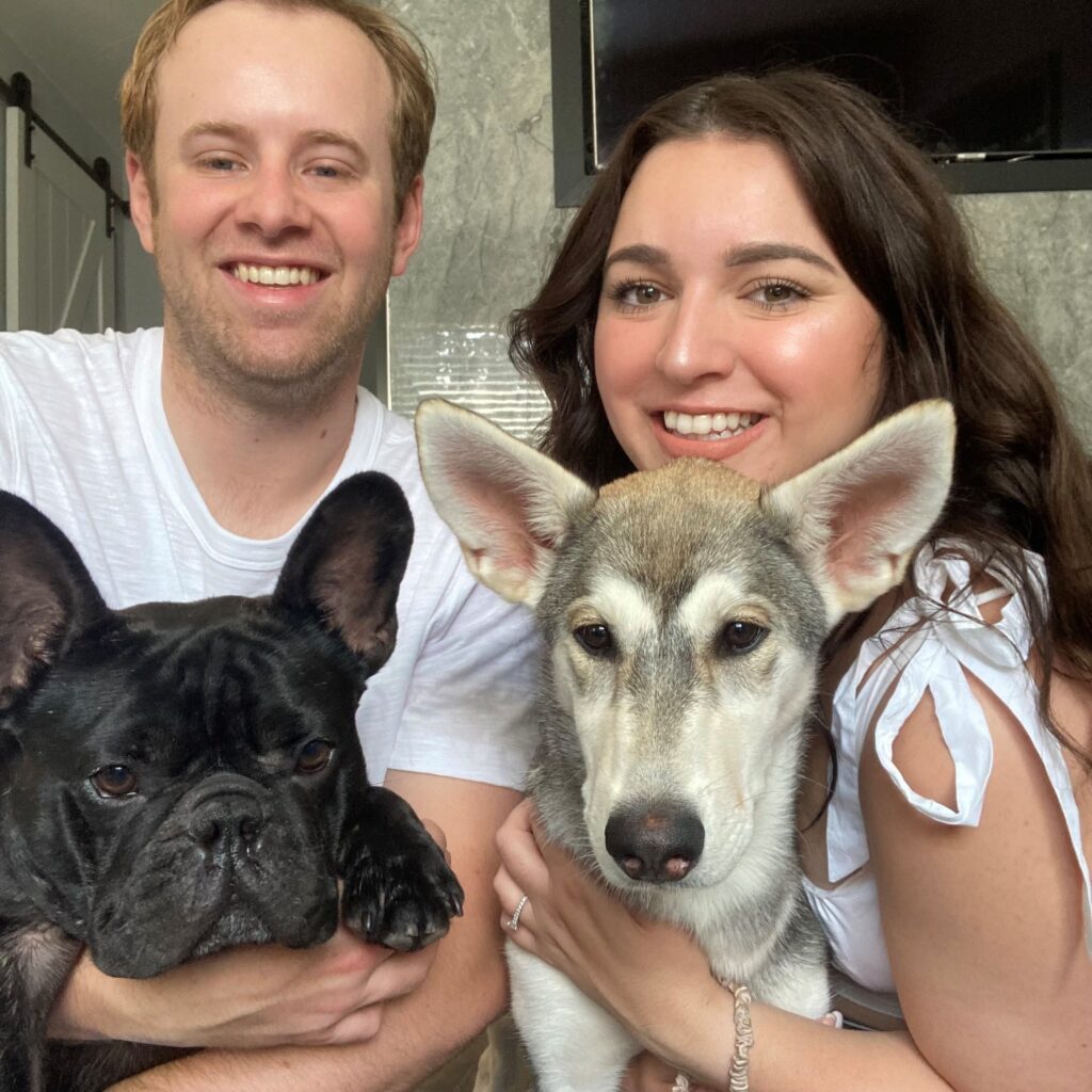 A smiling man and woman holding a black French bulldog and a grey and white husky mix, posing together indoors.