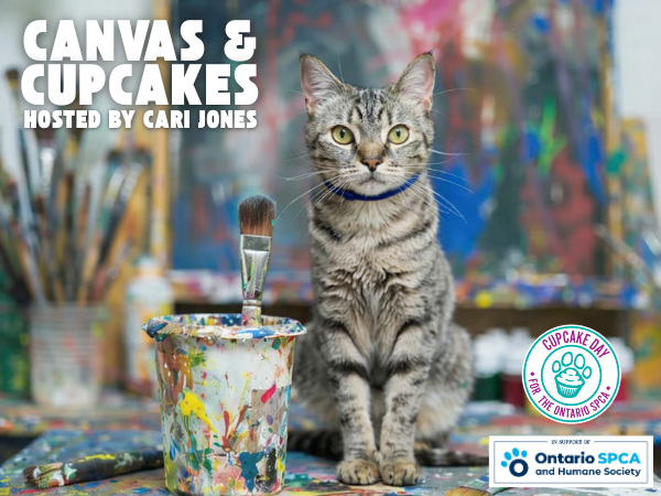 Image for Canvas and Cupcakes event. A gray tabby cat wearing a blue collar sits on a paint-splattered table in an art studio, facing the camera, with a paintbrush standing in a colorful cup and abstract paintings and brushes blurred in the background.