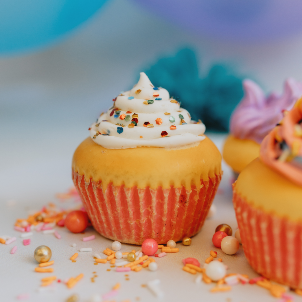 Close-up of a yellow-frosted cupcake in an orange wrapper topped with white icing and colorful sprinkles, with scattered candy decorations and blurred cupcakes in the background.