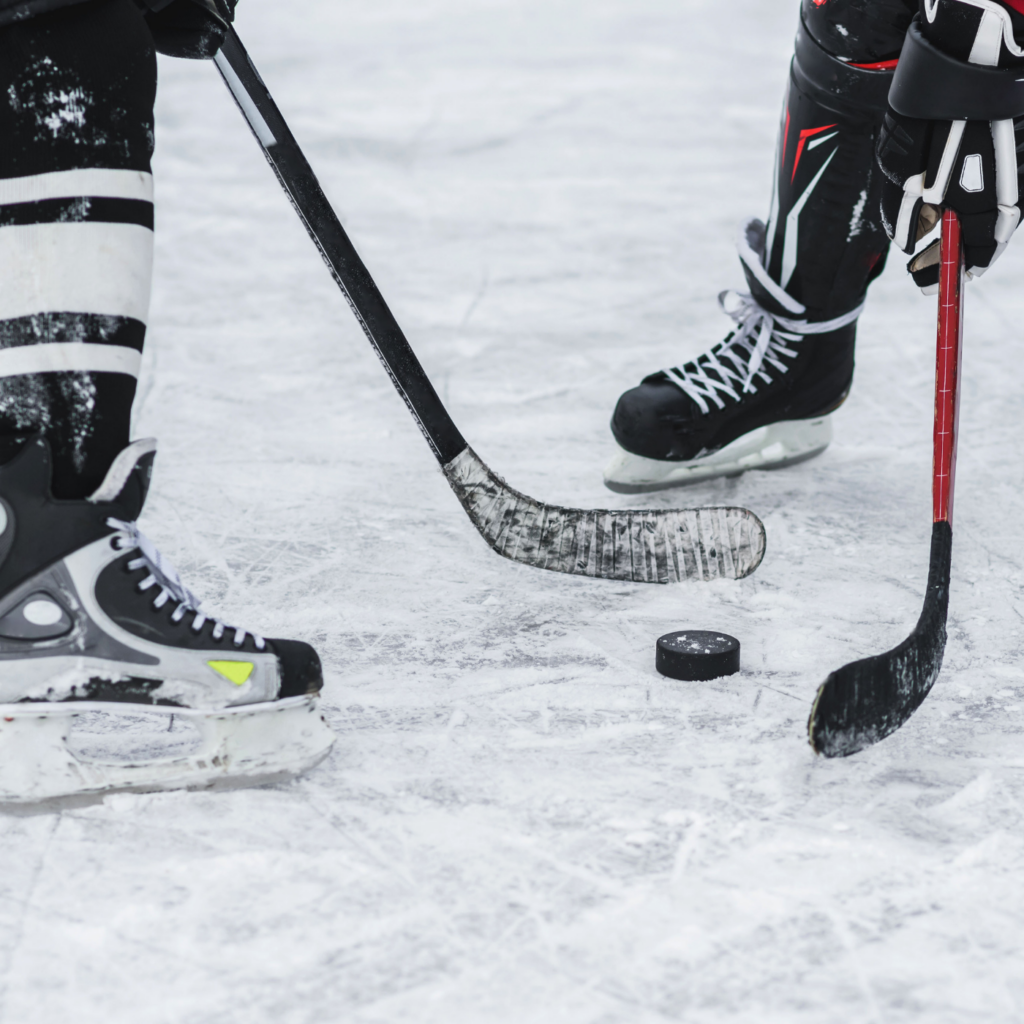 Close-up of two hockey players’ skates and sticks facing off over a puck on the ice, moments before a play begins.