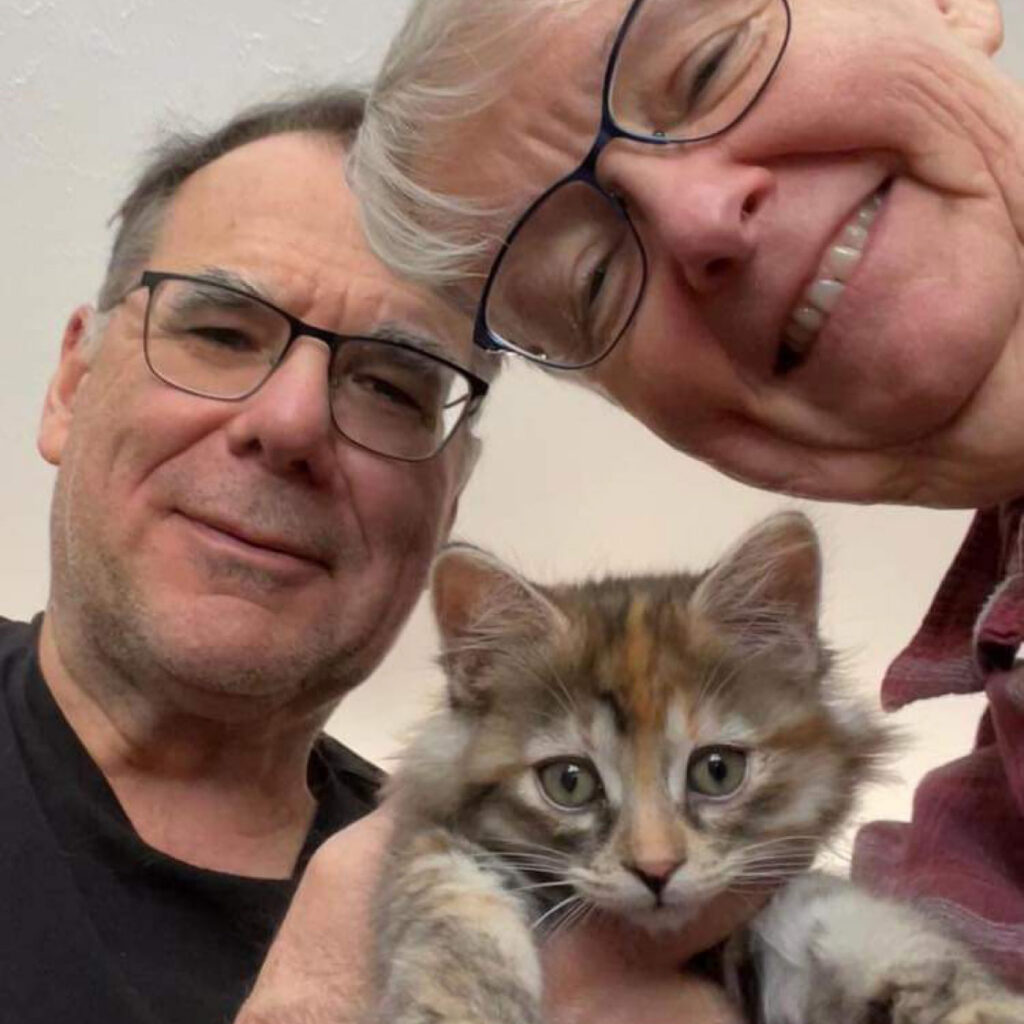 Close-up selfie of two adults smiling while holding a small calico kitten looking toward the camera.