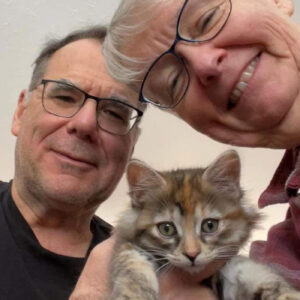 Close-up selfie of two adults smiling while holding a small calico kitten looking toward the camera.