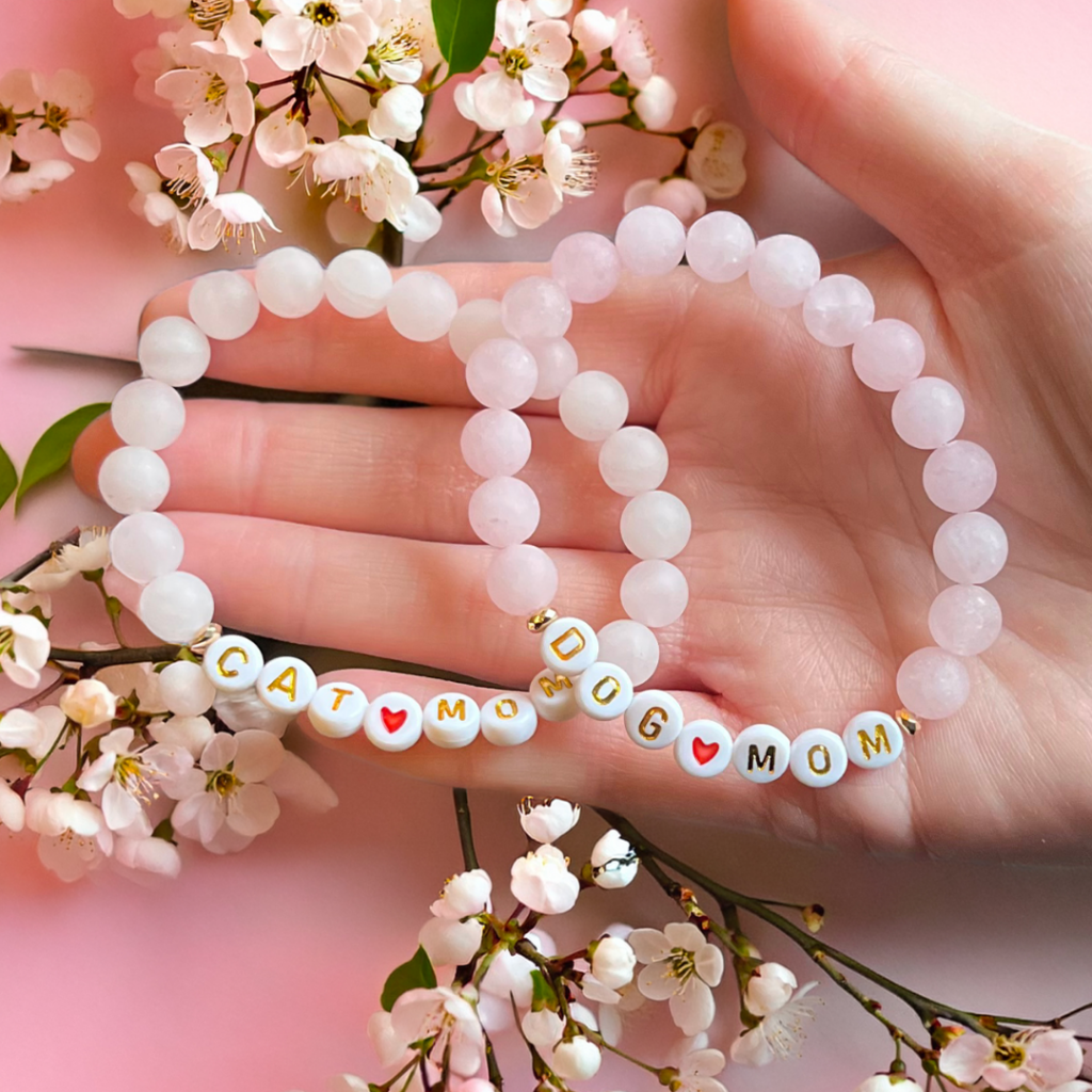 Hand holding “Cat Mom” and “Dog Mom” beaded bracelets with pink flowers in the background