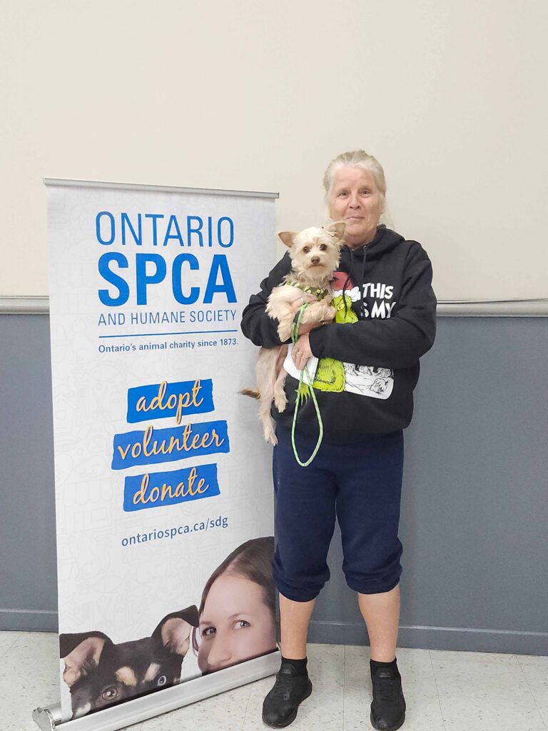 Woman holding a small tan dog beside an Ontario SPCA and Humane Society banner promoting adoption, volunteering, and donations at a community pet event.