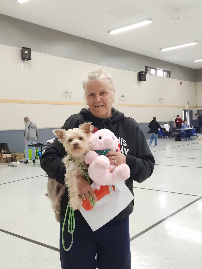Smiling woman holding a small tan dog and a pink stuffed pig toy inside a large community hall during an Ontario SPCA pet event.
