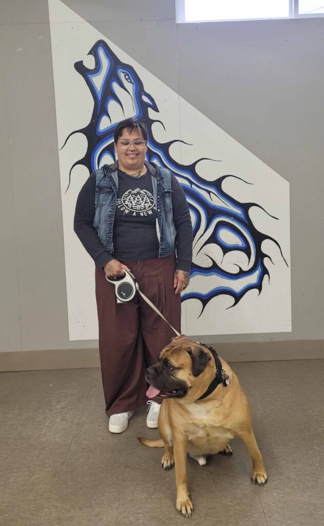 Person standing with a large tan mastiff-type dog on a leash in front of a blue, white, and black Indigenous-style mural.