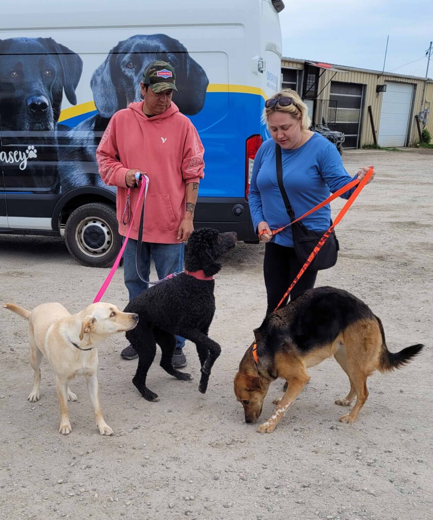 Two people with three dogs gathered beside a transport van during an animal transfer or community pet event.