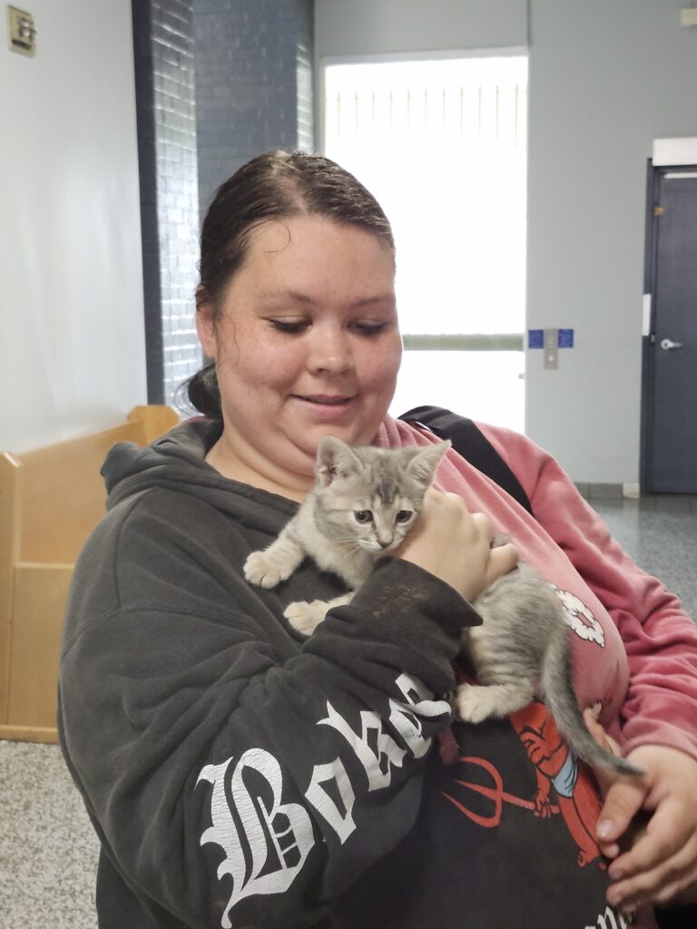 Person holding a small gray tabby kitten indoors while smiling down at it.