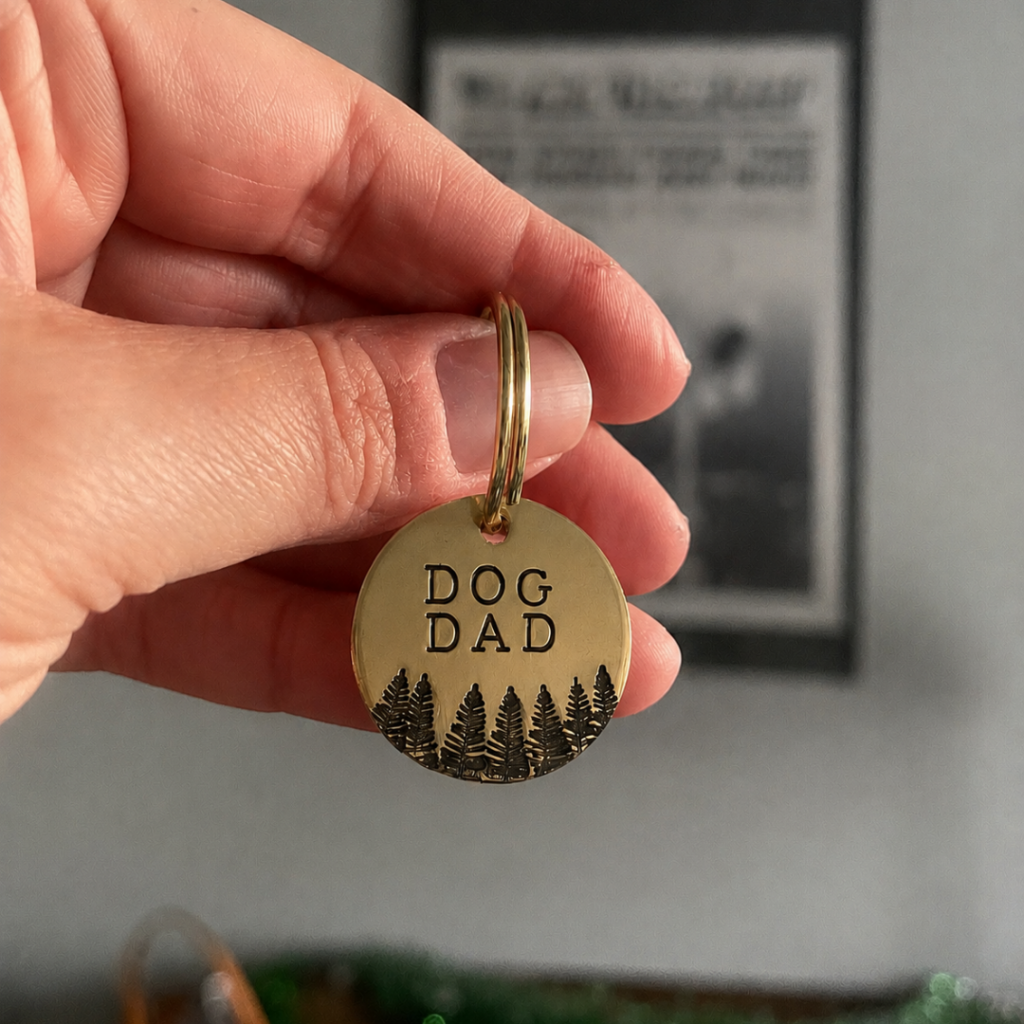 A close-up shot of a hand holding a round, polished gold-toned metal keychain tag. The text 'DOG DAD' is stamped in the center in a simple, dark font, with an engraved graphic of a forest of pine trees along the bottom edge of the tag.