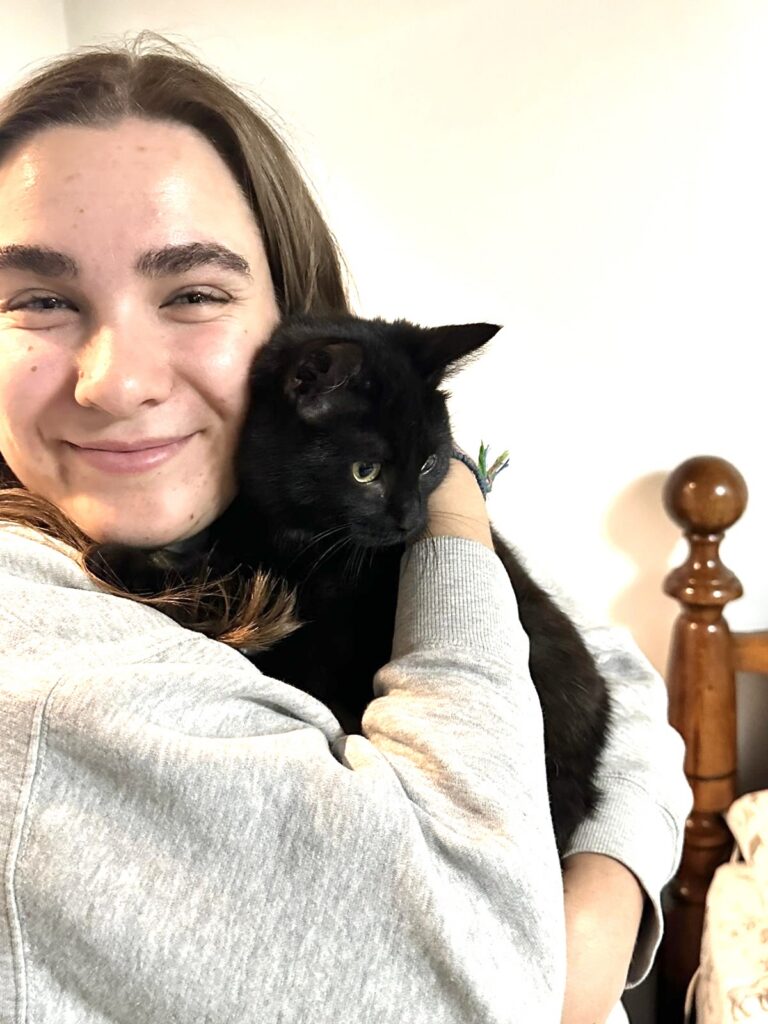 A girl smiling while holding a black cat close to their face in a sunlit room.