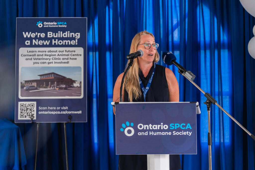 A woman speaks at a podium during an Ontario SPCA and Humane Society event with a display board behind her announcing plans for a new animal centre and veterinary clinic, with blue curtains forming the backdrop.