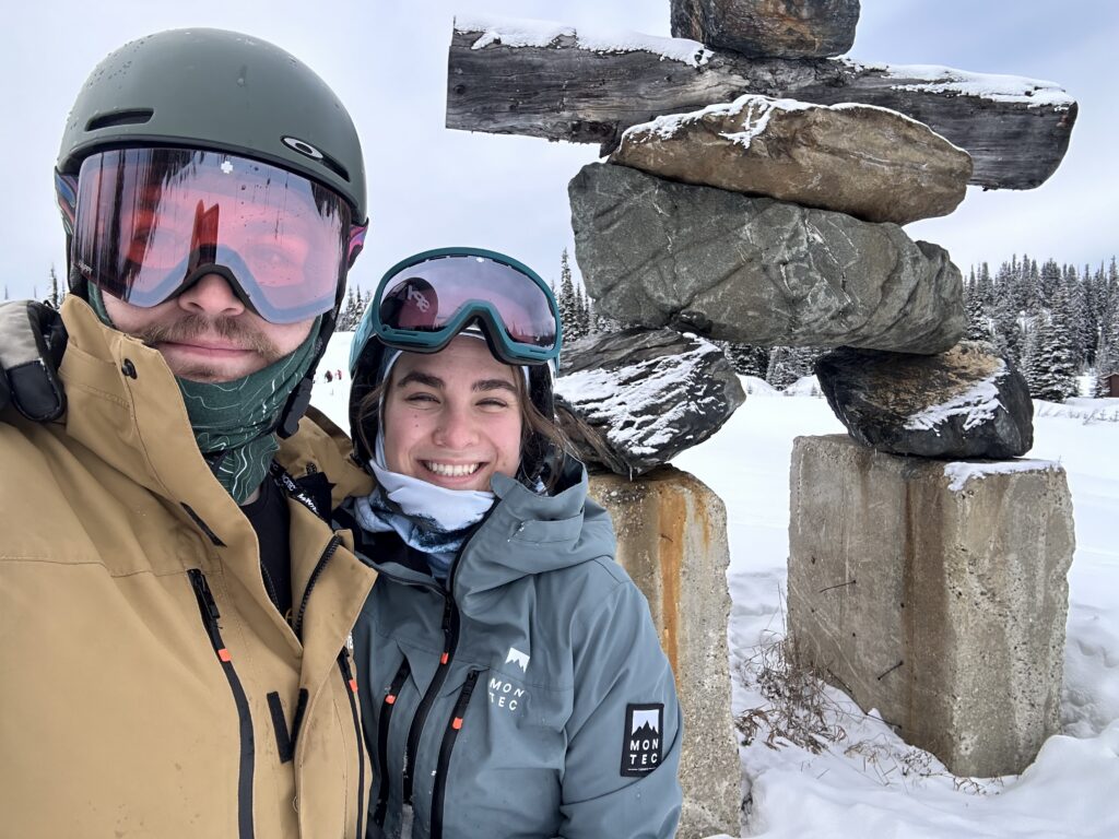 Two people in ski gear pose for a selfie beside a traditional stone inukshuk on a snowy mountain.