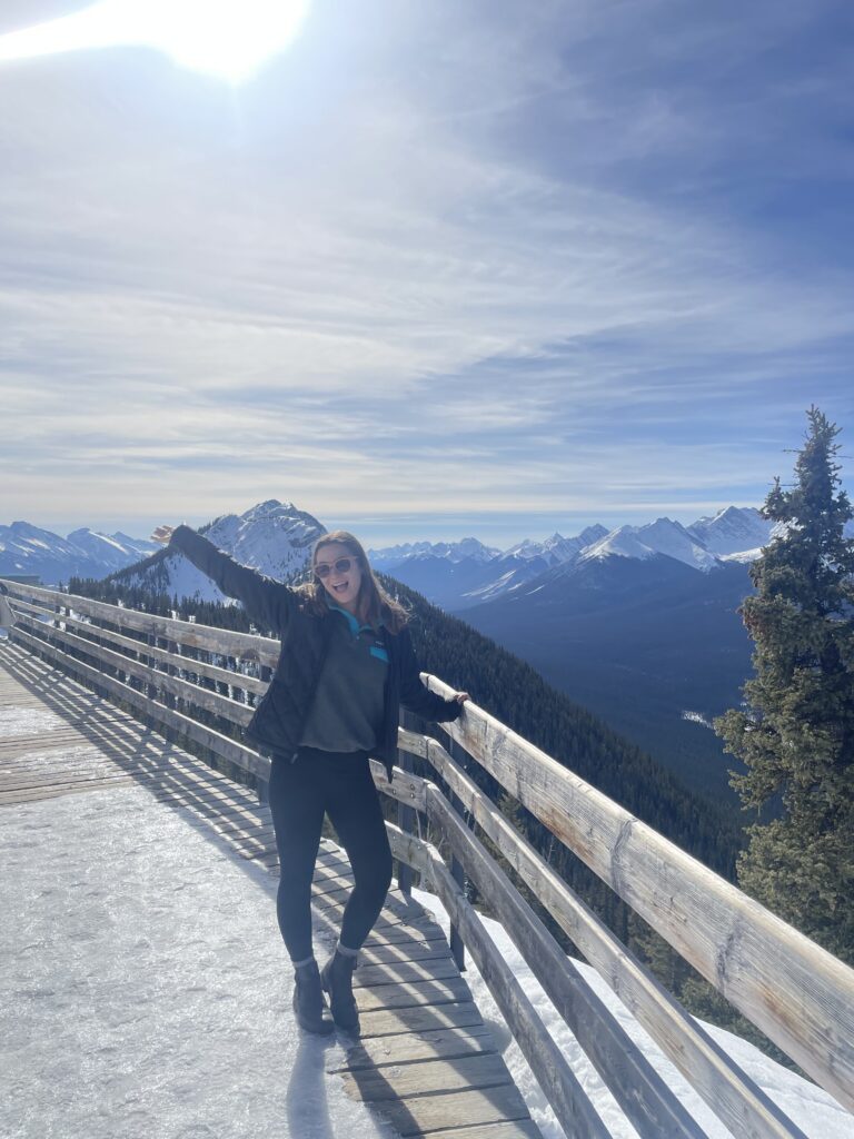 A girl poses on a snowy mountain lookout with panoramic views of rugged peaks and forested valleys.