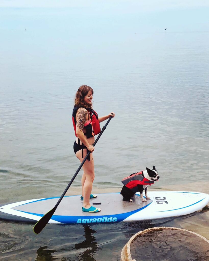 Person and small dog wearing life jackets on a paddleboard in calm lake water.