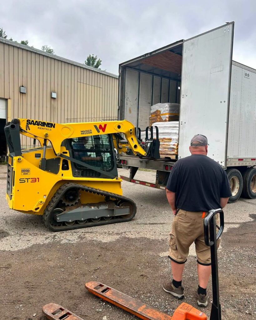 A skid steer loader transfers pallets of pet food into the back of a large transport truck while a worker watches nearby in an industrial loading area.