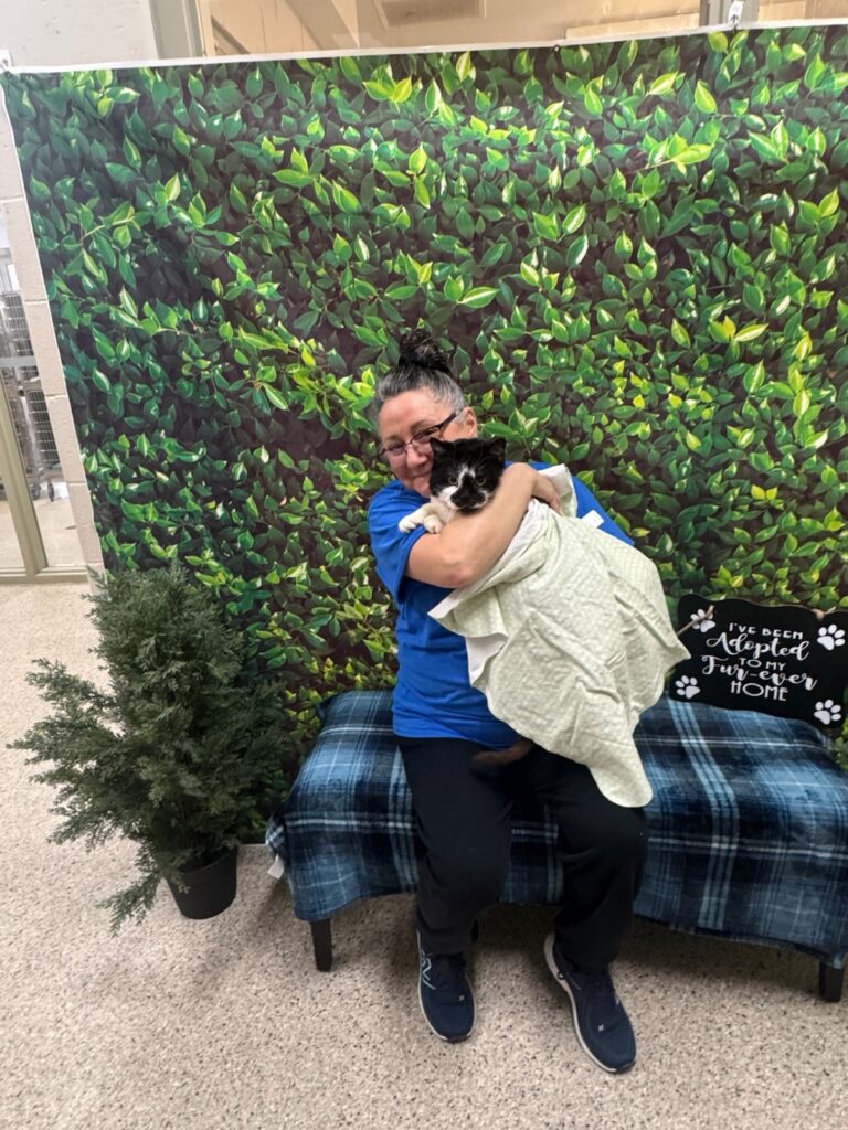 A woman cuddles black-and-white cat after his adoption at an animal shelter, sitting in front of a green leafy backdrop with an "I've Been Adopted to My Furever Home" sign.