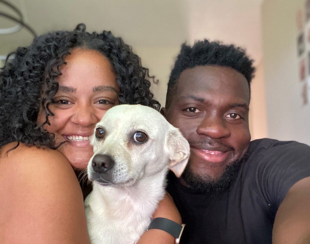 Smiling couple cuddles a small white dog close between them in a selfie at home