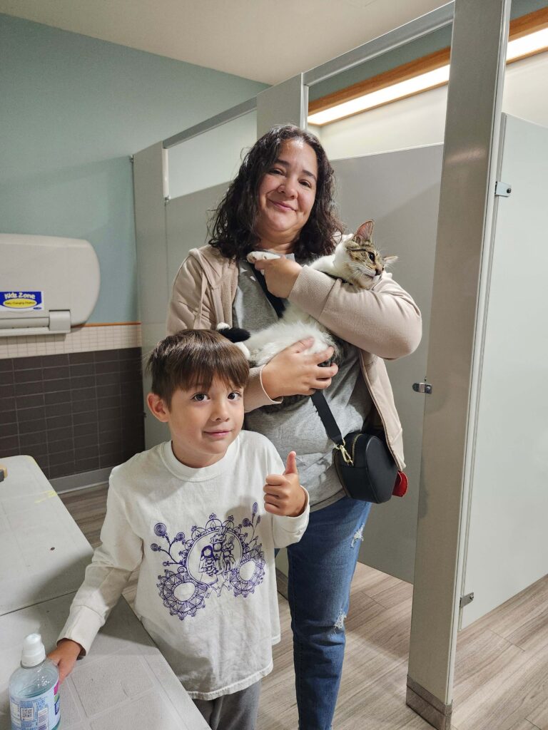 Woman and young child holding tabby and white kitten in shelter facility bathroom with child giving thumbs up