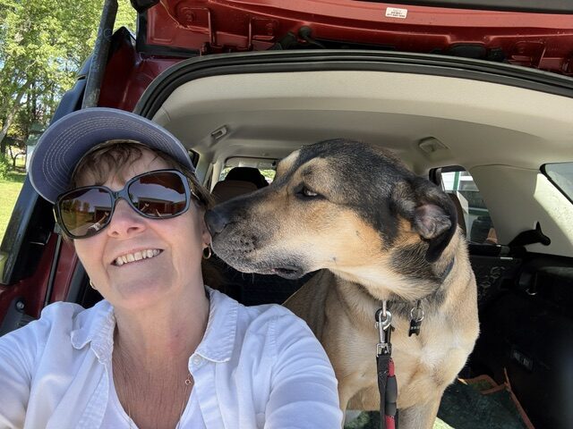 A woman smiles for a selfie in the open trunk of an SUV as a tan and black dog leans in close beside her.