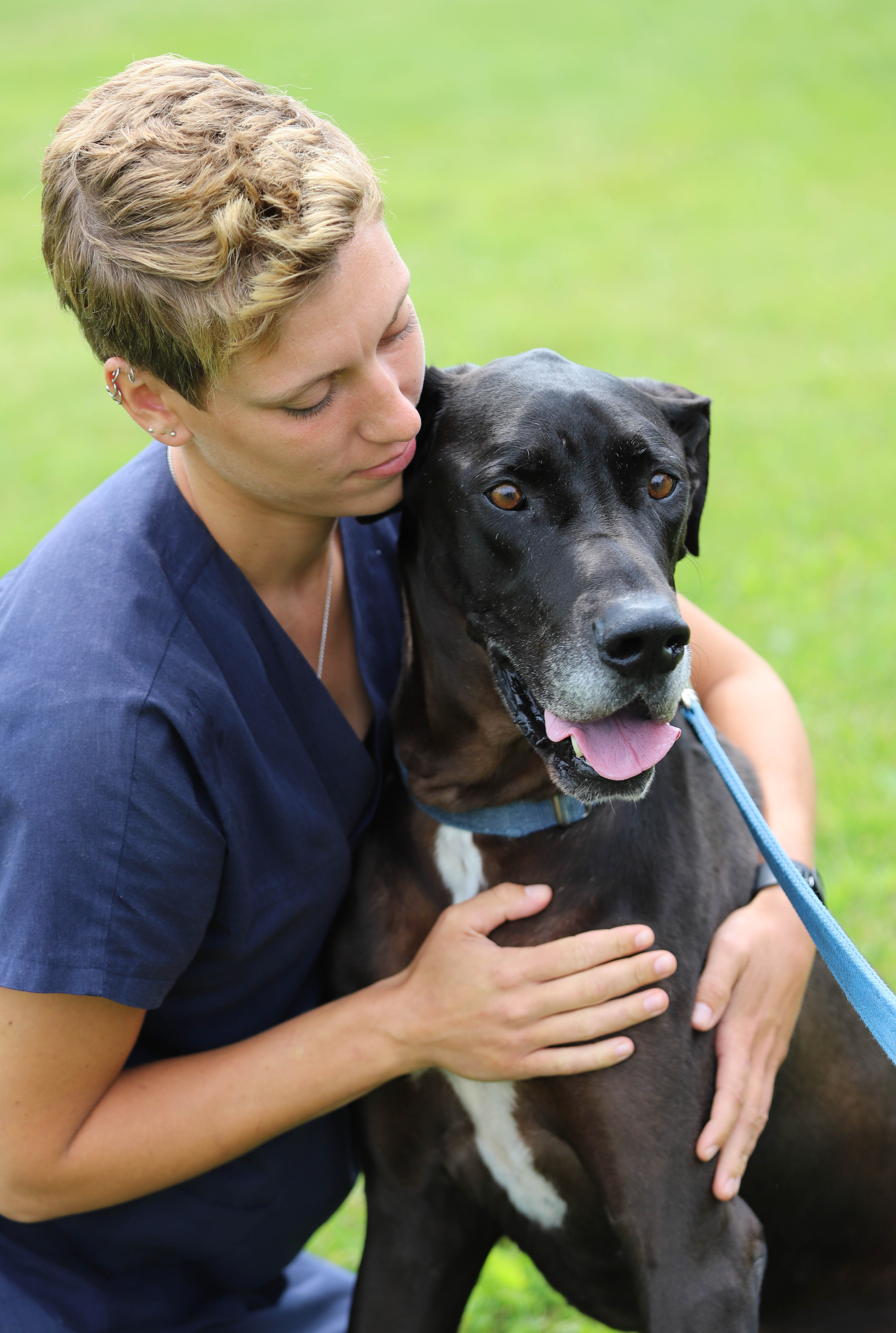 Megan Holmes in navy scrubs resting her cheek against a large black dog with a greying muzzle, one arm around the dog, on a grassy lawn