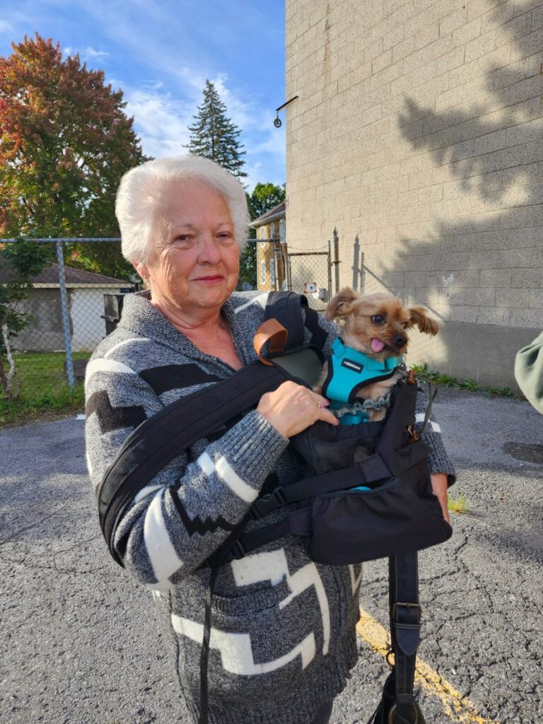 A smiling woman with white hair carries a small long-haired dog in a teal harness in a front sling.
