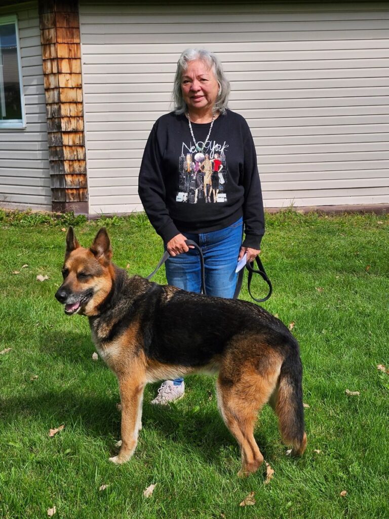 A woman with silver hair stands on the grass holding the leash of a large tan and black dog.