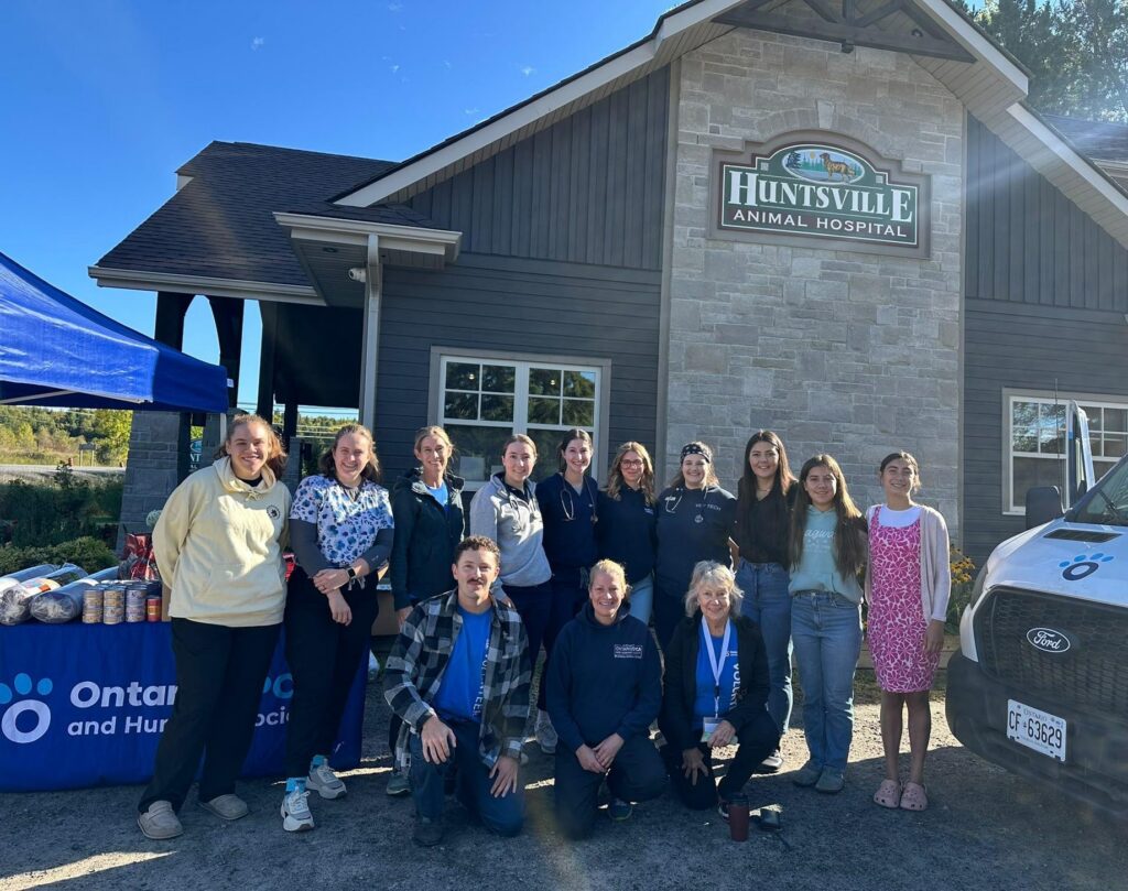 Thirteen people pose outside Huntsville Animal Hospital beside an Ontario SPCA tent, a table of pet food and a van.