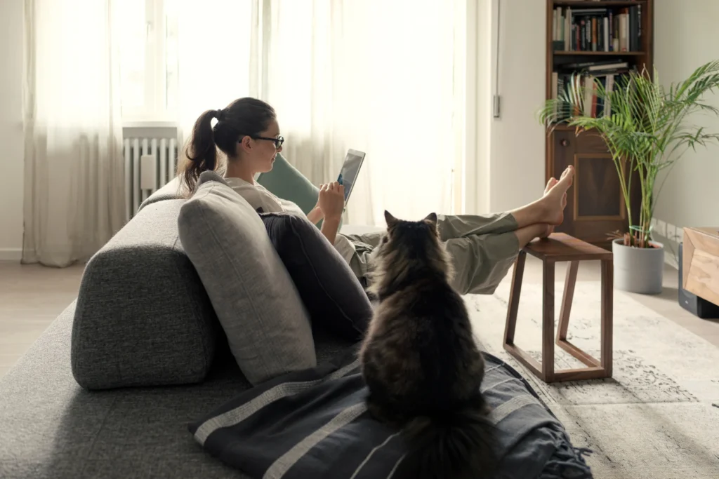 Woman wearing glasses sitting on floor with black cat while using tablet in bright modern living room