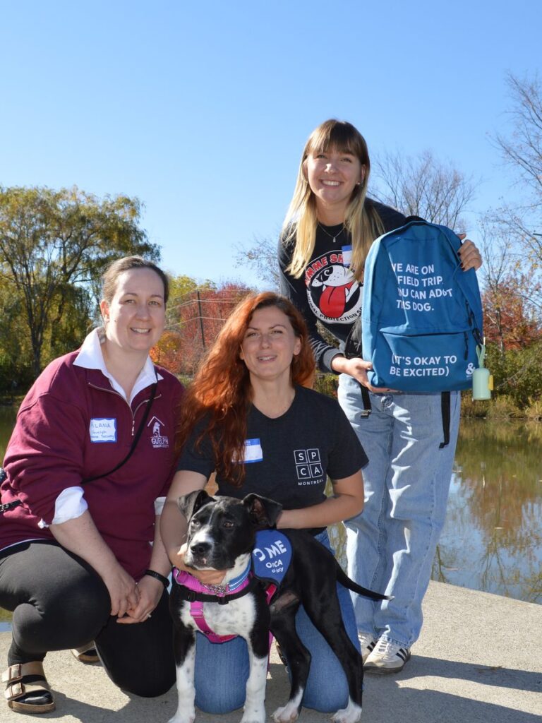Three people by a pond with a black and white puppy, one holding a backpack that reads We are on a field trip